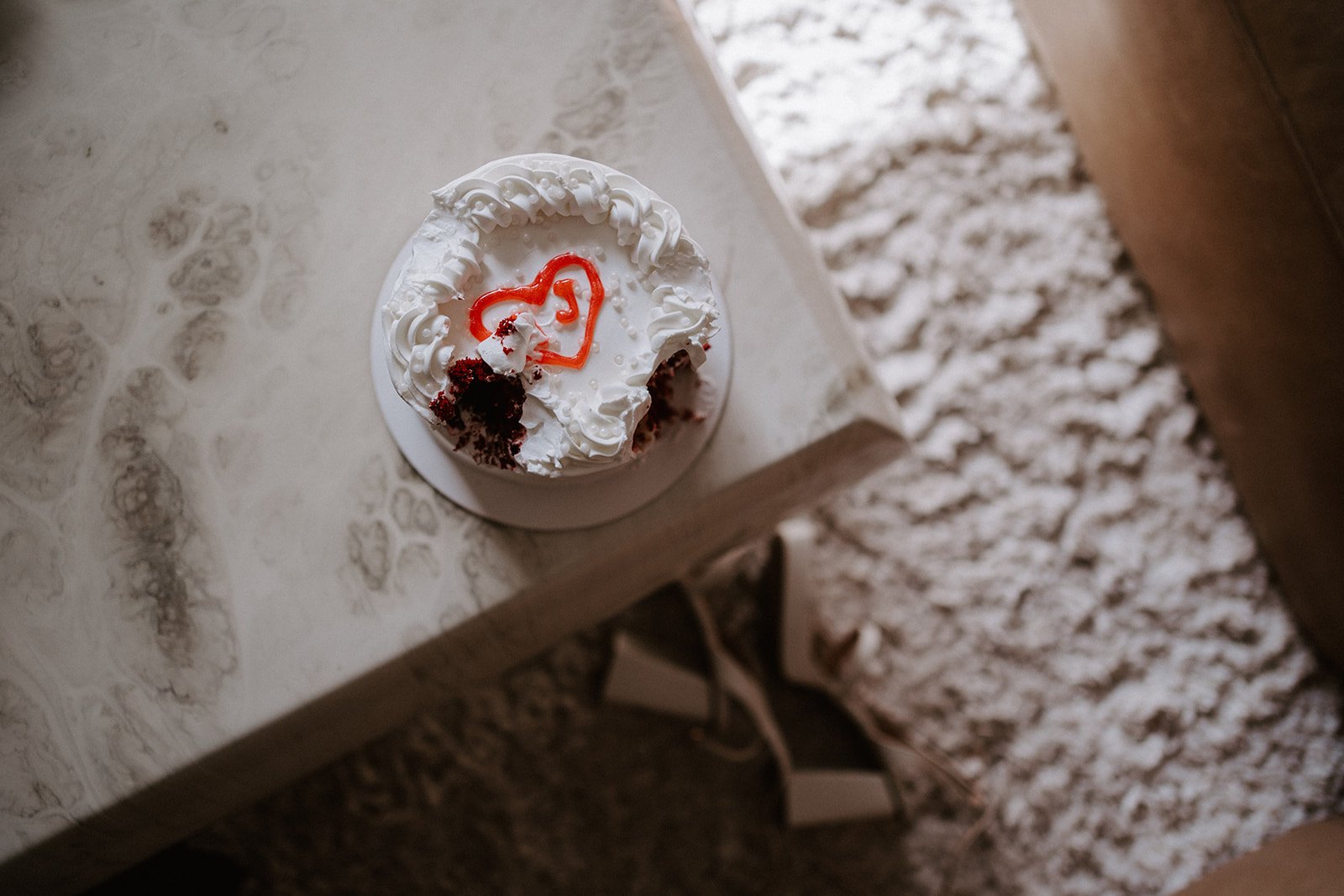 Small cake with white frosting and red heart decoration, partially eaten, on a marble-like table surface.