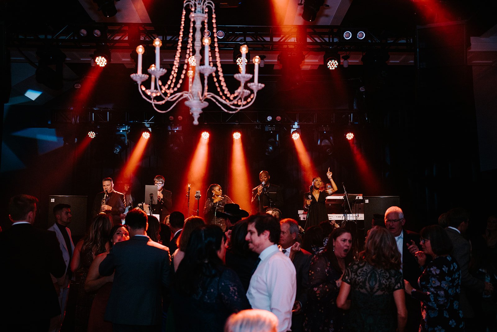 A live band performs on stage at an indoor event, with a chandelier overhead and people dancing in front. Red stage lights illuminate the area, creating a vibrant atmosphere.