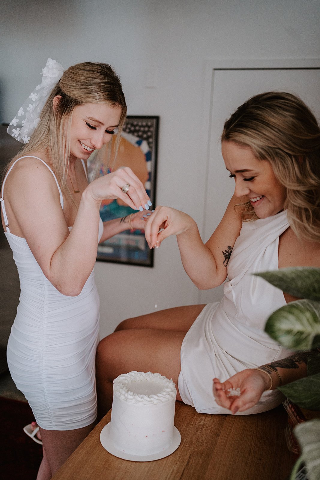 Two women in white dresses smiling and interacting near a small white cake on a table, with one holding a handful of sprinkles. A plant is visible in the foreground.