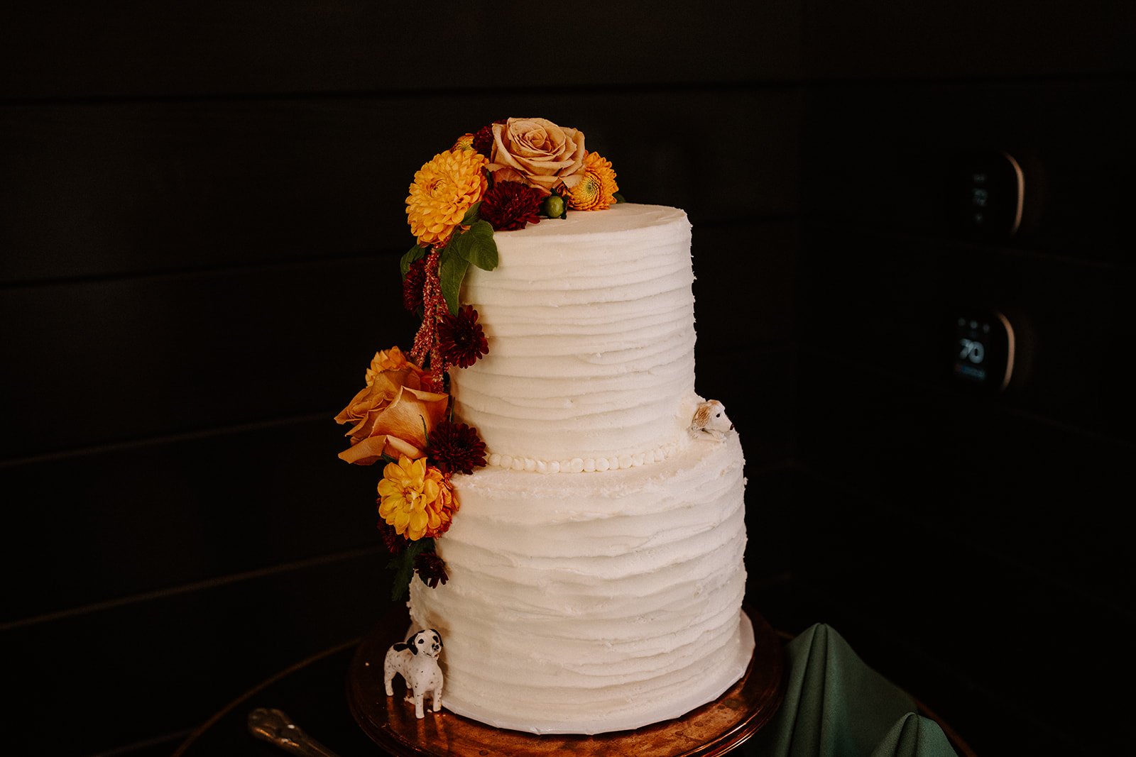 Two-tier white cake with textured frosting, decorated with orange and yellow flowers, and small animal figurines on a wooden cake stand.