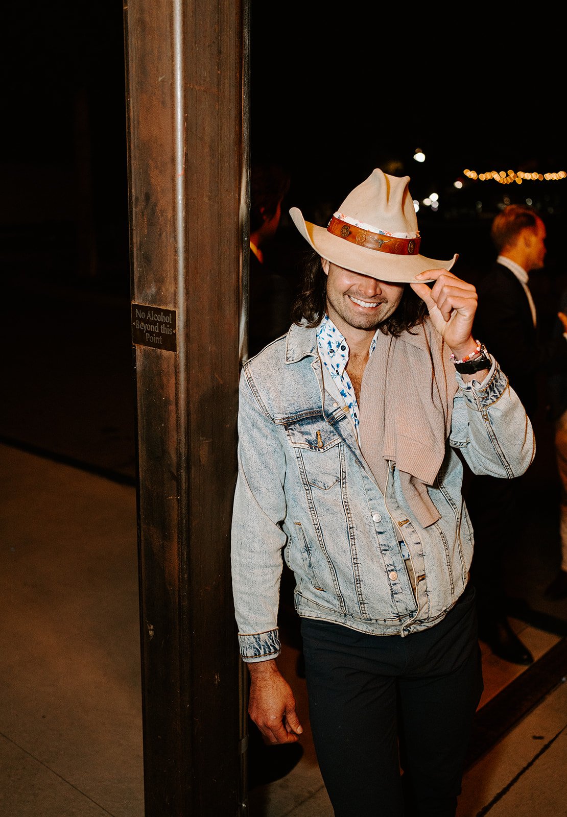 Smiling person in a cowboy hat and denim jacket leaning against a post with string lights in the background.