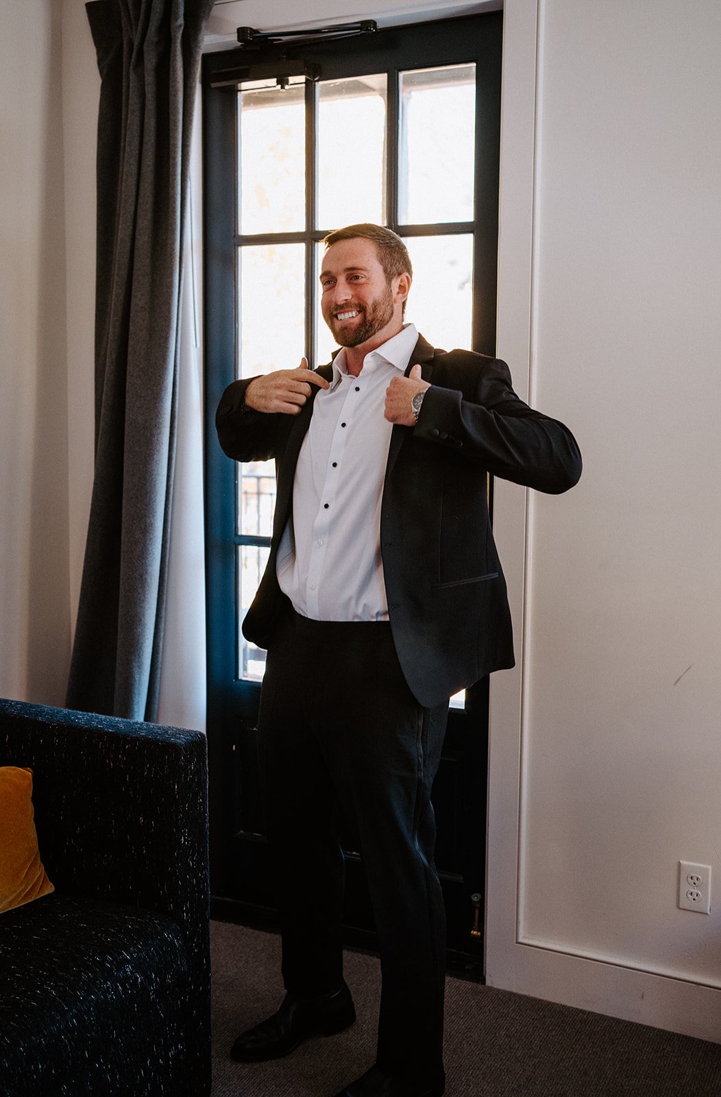 A man in a black suit and white shirt, smiling and adjusting his collar, stands near a window in an indoor setting.