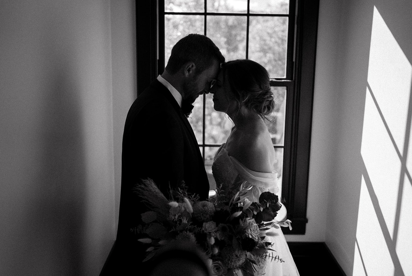 Silhouette of a couple embracing with their foreheads touching in front of a window, woman holding a bouquet, black and white.