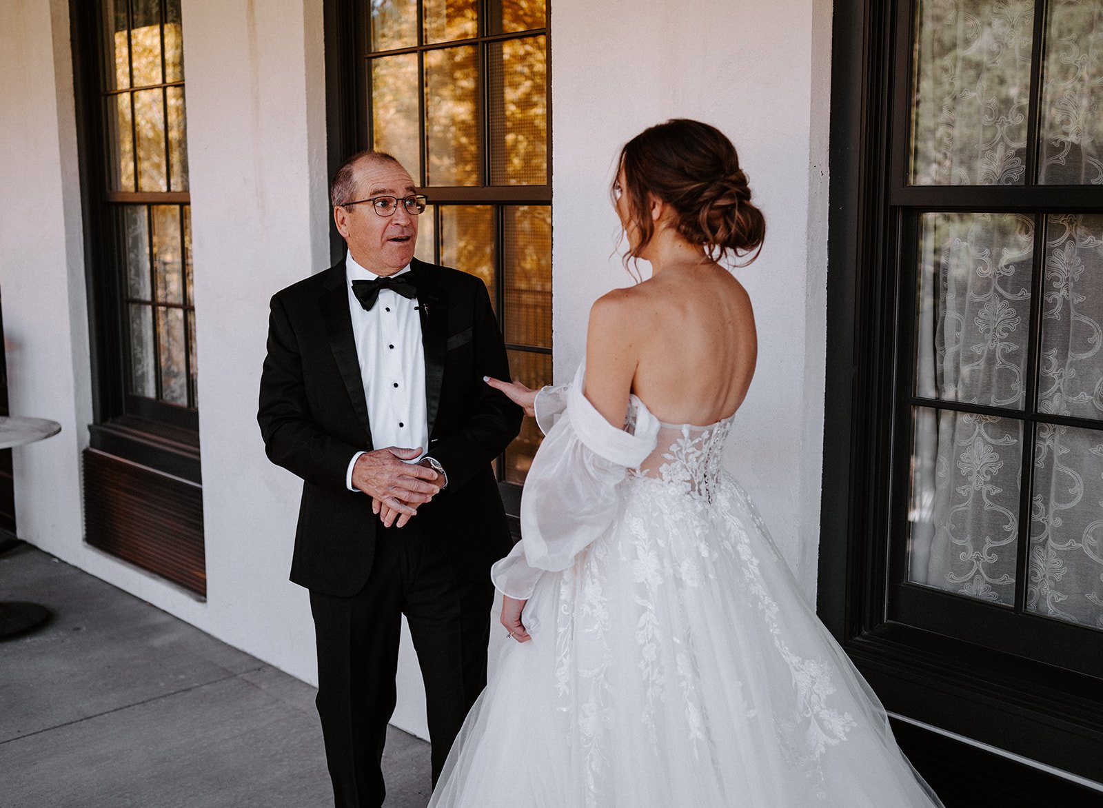 A bride in a white wedding dress and a man in a tuxedo stand outside near a window.
