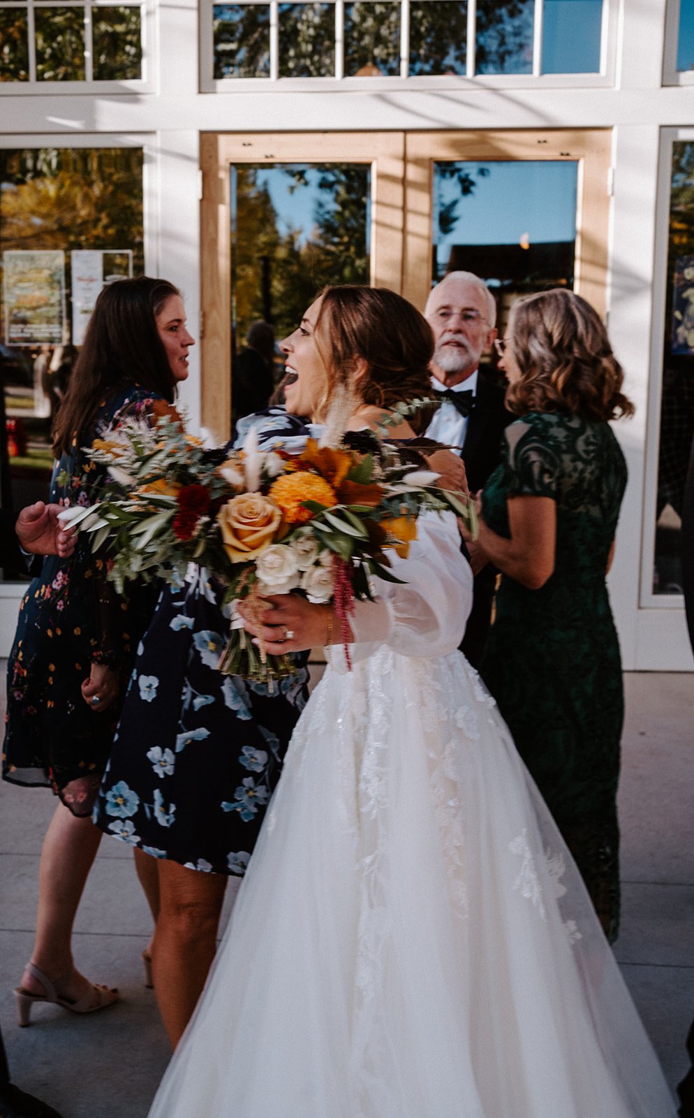 Bride hugging a guest with a bouquet in hand, surrounded by people in formal attire, outside a building with glass doors.