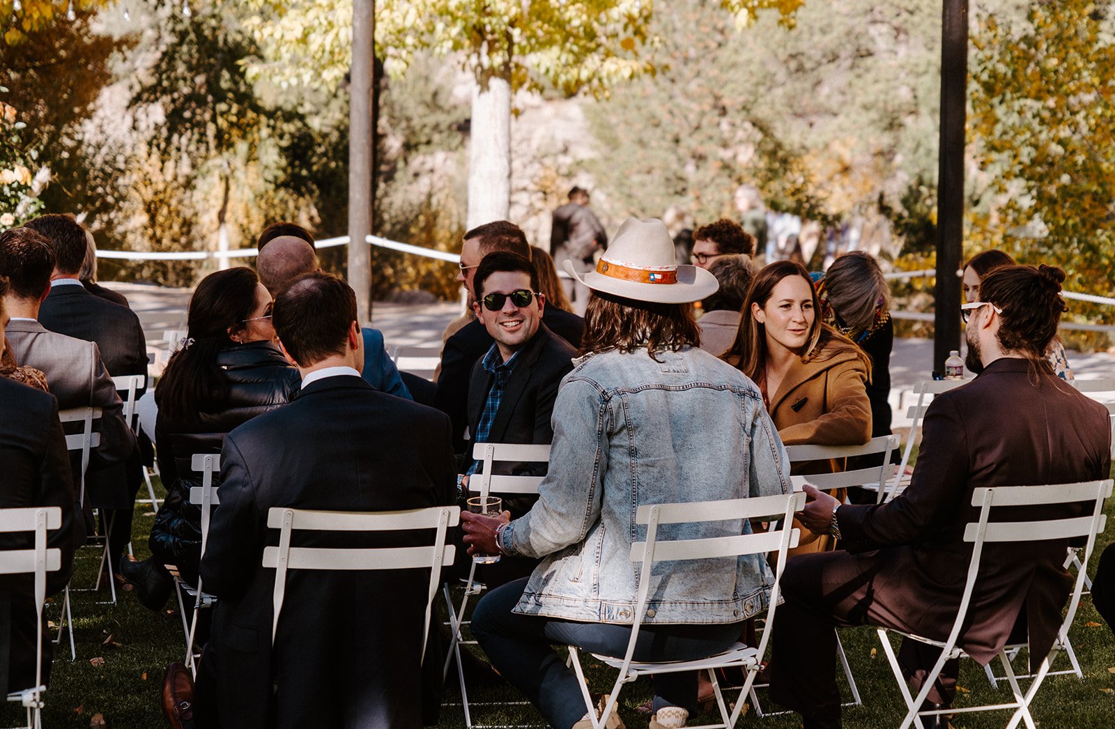Outdoor event with people seated on white chairs, socializing in an autumn setting with trees in the background.