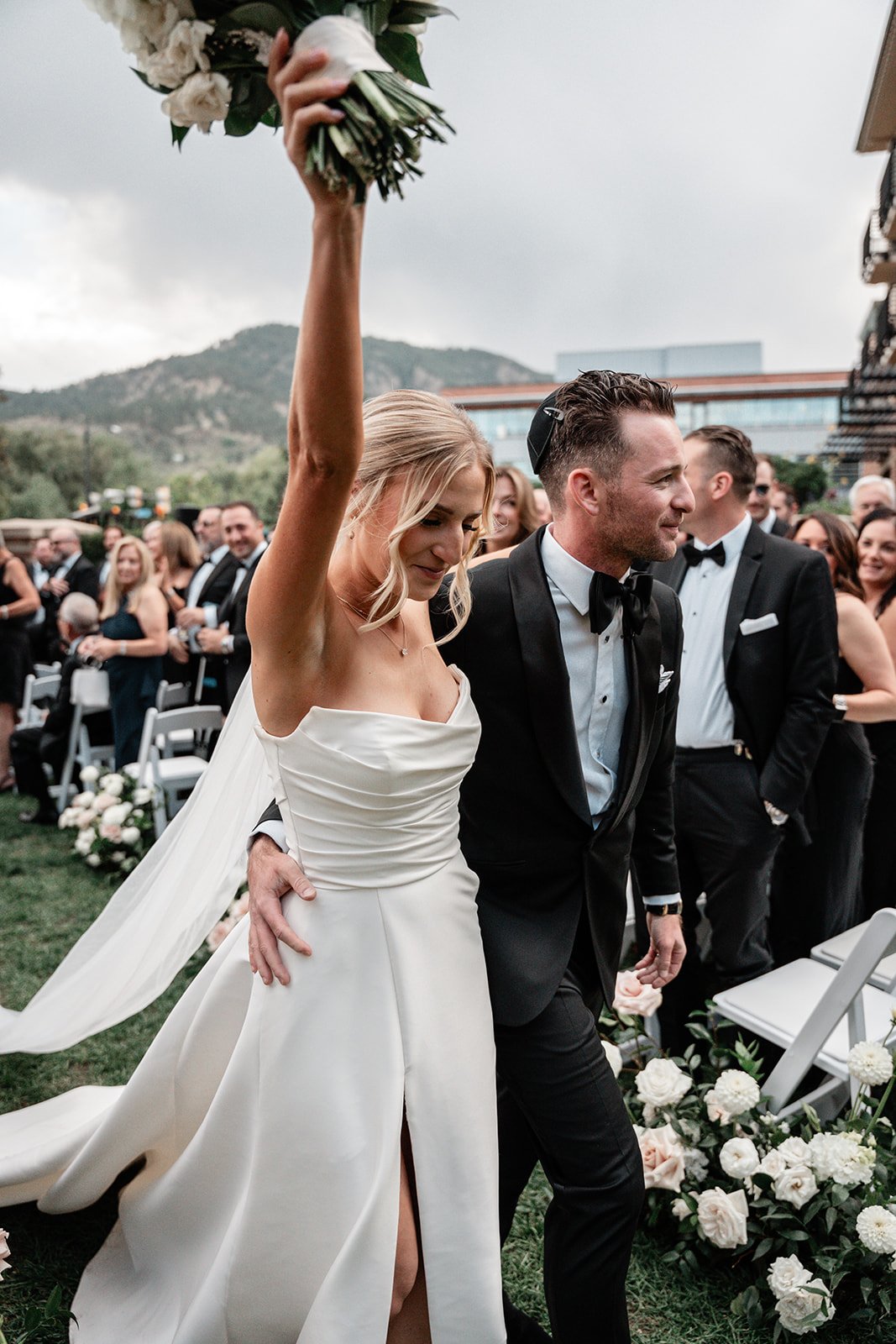 Bride and groom celebrating at outdoor wedding ceremony, surrounded by guests, with bride holding a bouquet.
