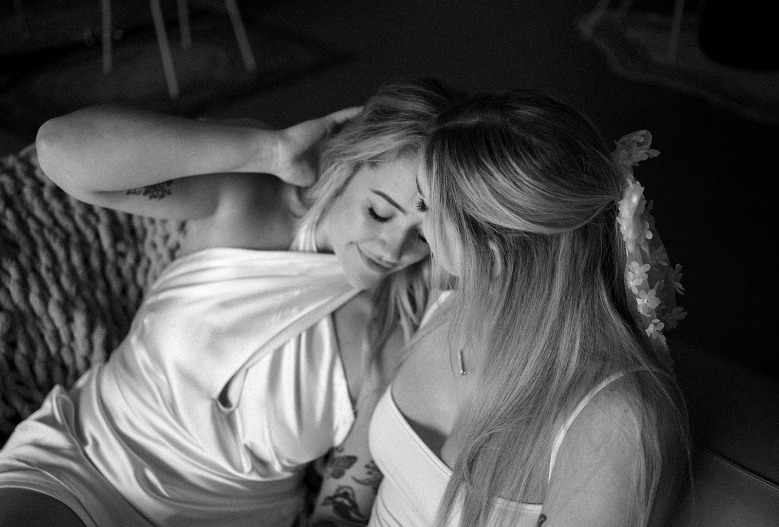 Two women sitting closely on a sofa, smiling and leaning towards each other, wearing white outfits, in a dimly lit room.