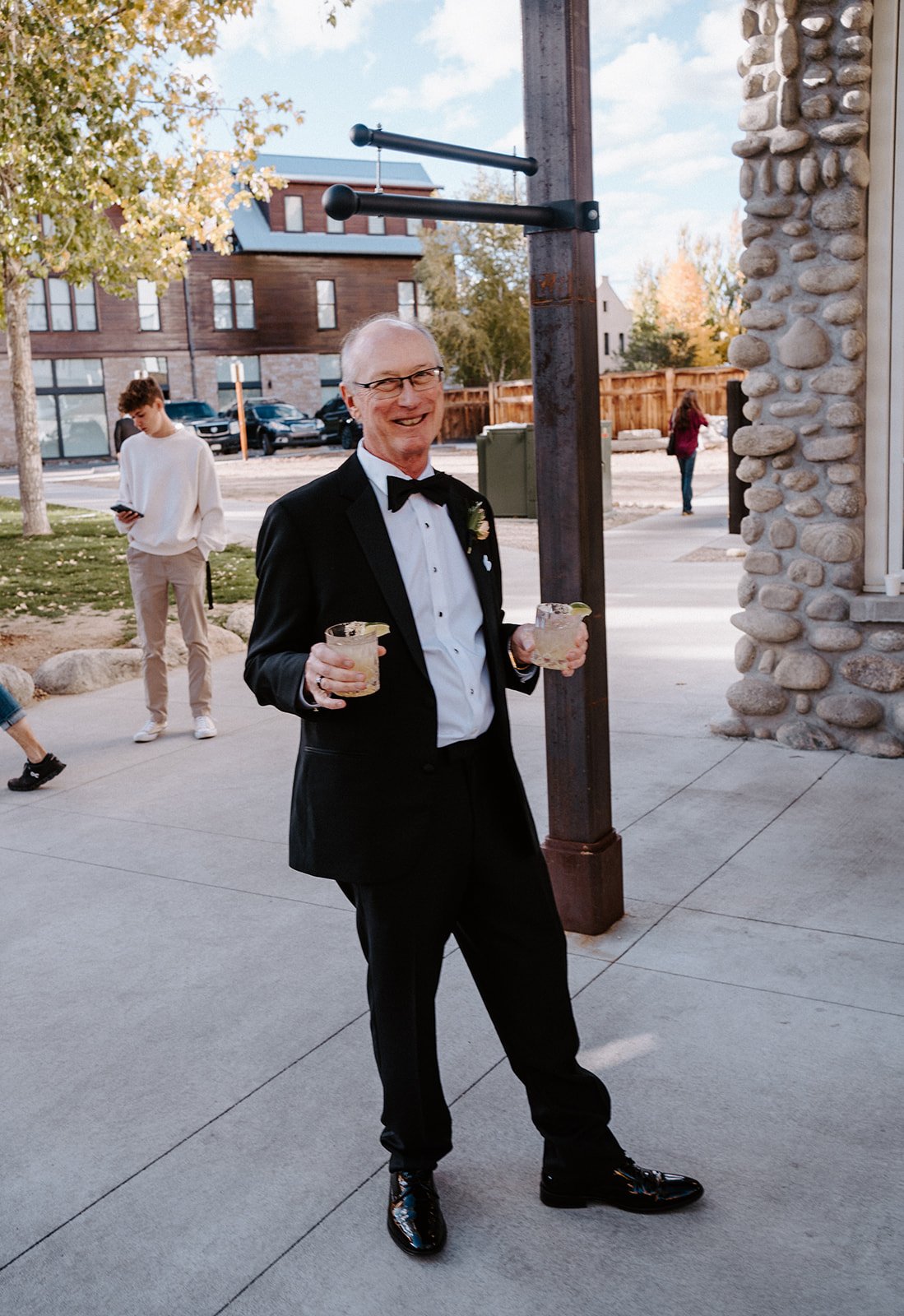 Man in a tuxedo holding drinks outdoors near a building with stone facade.