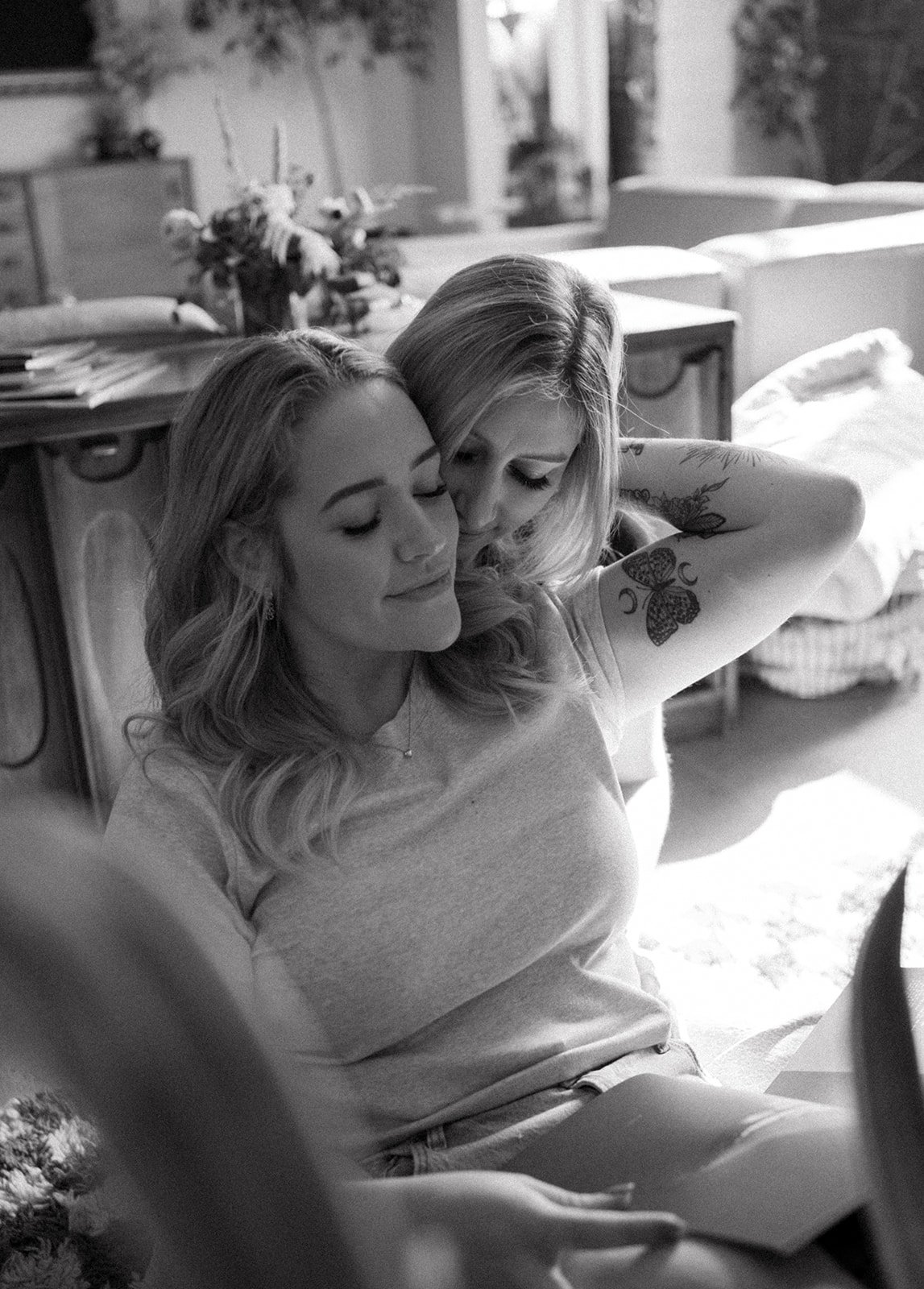 Two women sitting closely, one with a tattooed arm, appearing affectionate and content in a cozy indoor setting, captured in black and white.