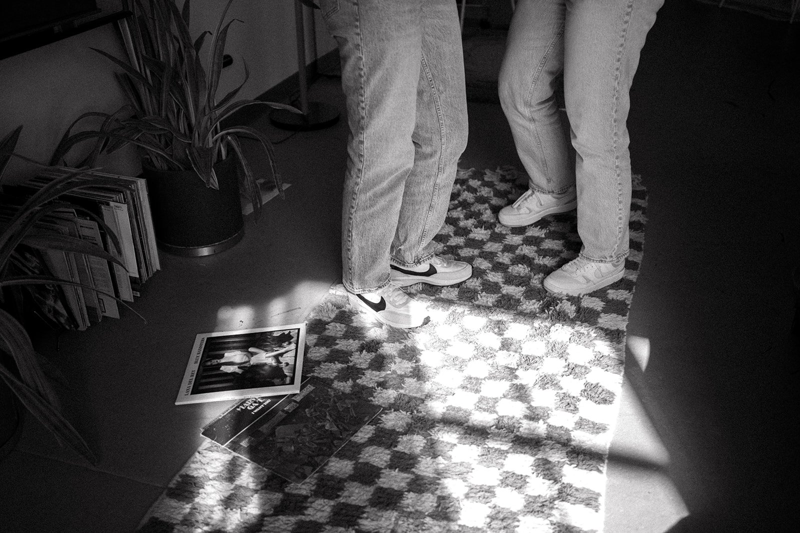 Black and white photo of two people standing on a checkered rug, wearing jeans and sneakers. A plant and a stack of magazines or books are nearby, with two photographs on the floor.
