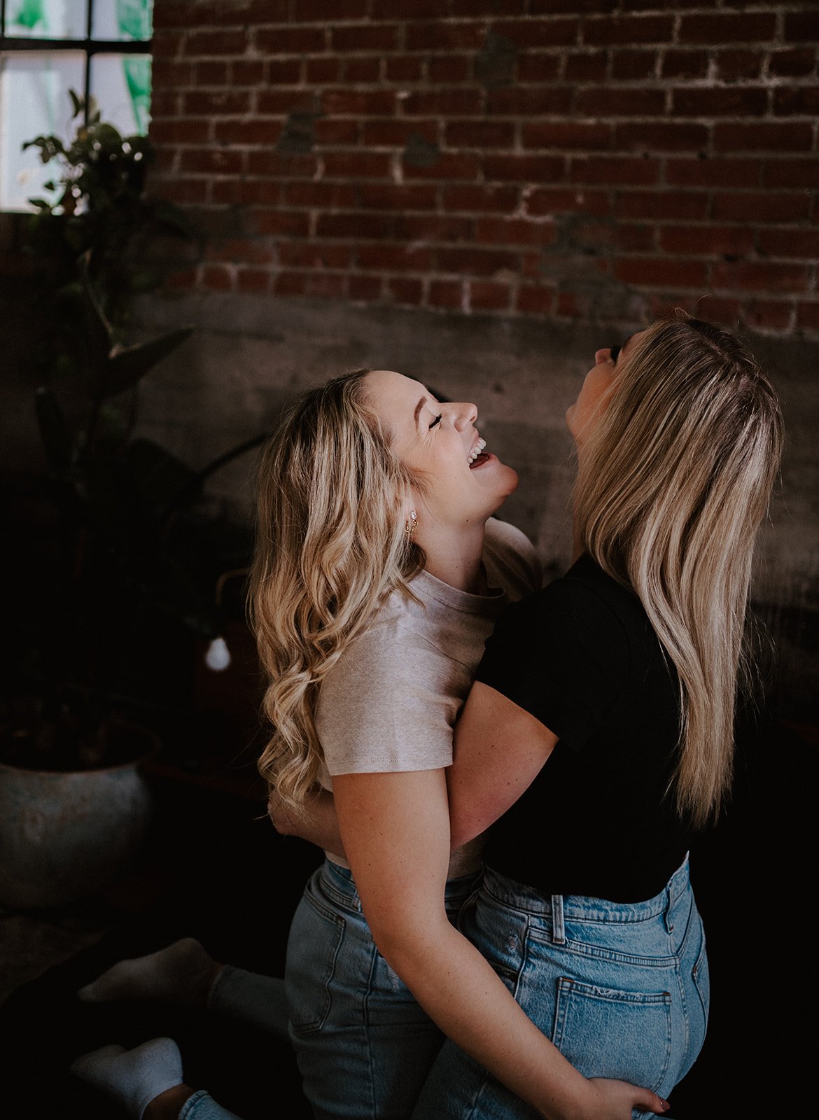 Two women laughing and hugging in front of a brick wall, wearing casual jeans and shirts.