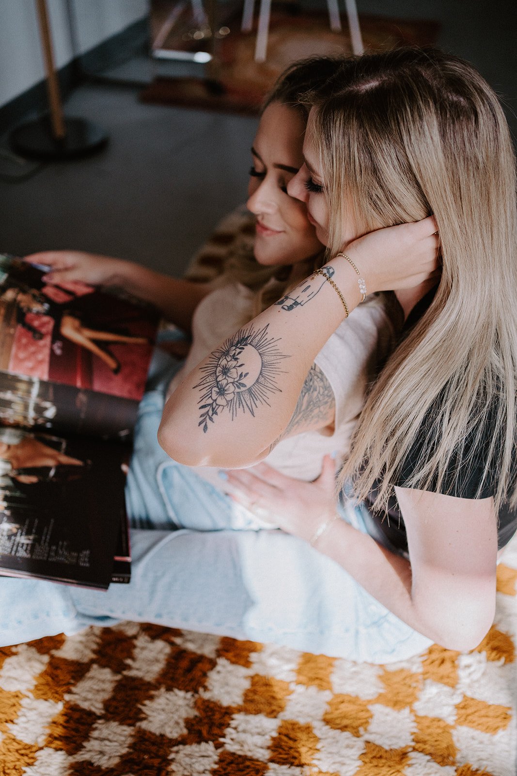 Two people sitting closely, one with a prominent arm tattoo, looking at an open magazine, on a checkered rug.