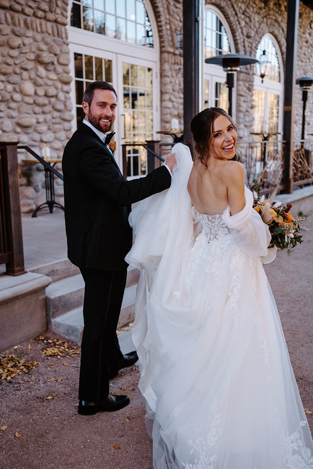 Bride and groom smiling outside a stone building, with the groom holding the bride's dress train, and the bride holding a bouquet.