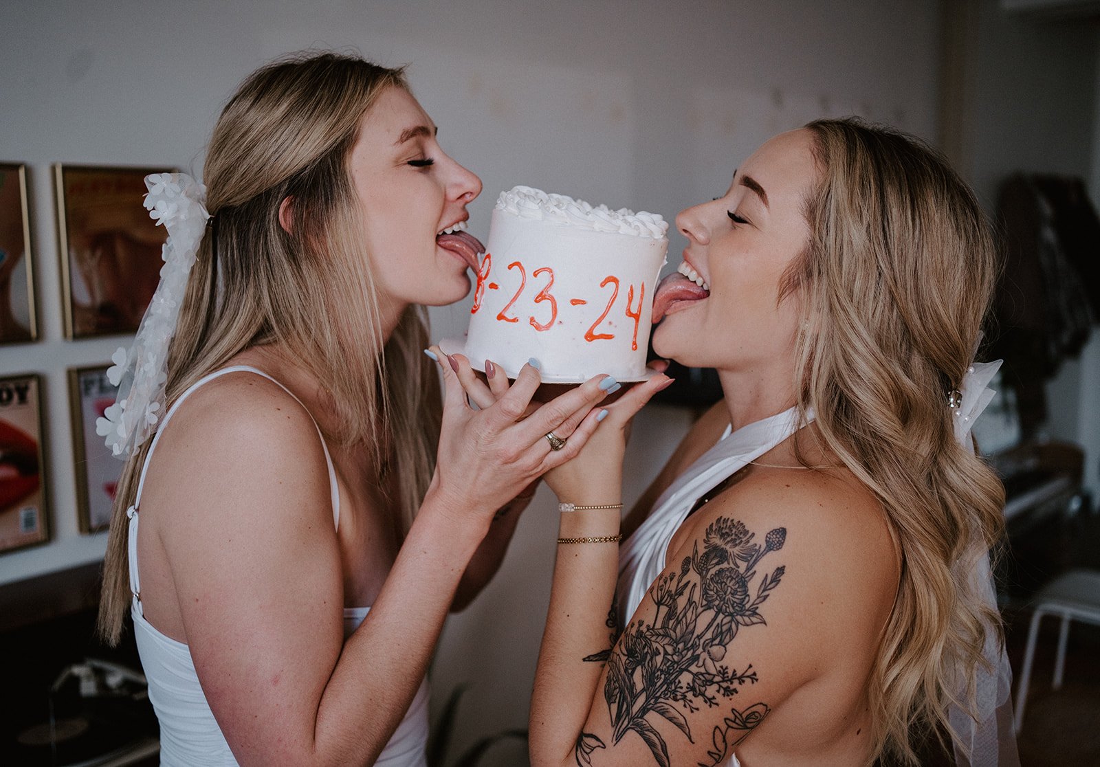 Two women with long hair and floral veils playfully licking a white cake with red-orange writing "3-23-24." One woman has a black floral tattoo on her arm.