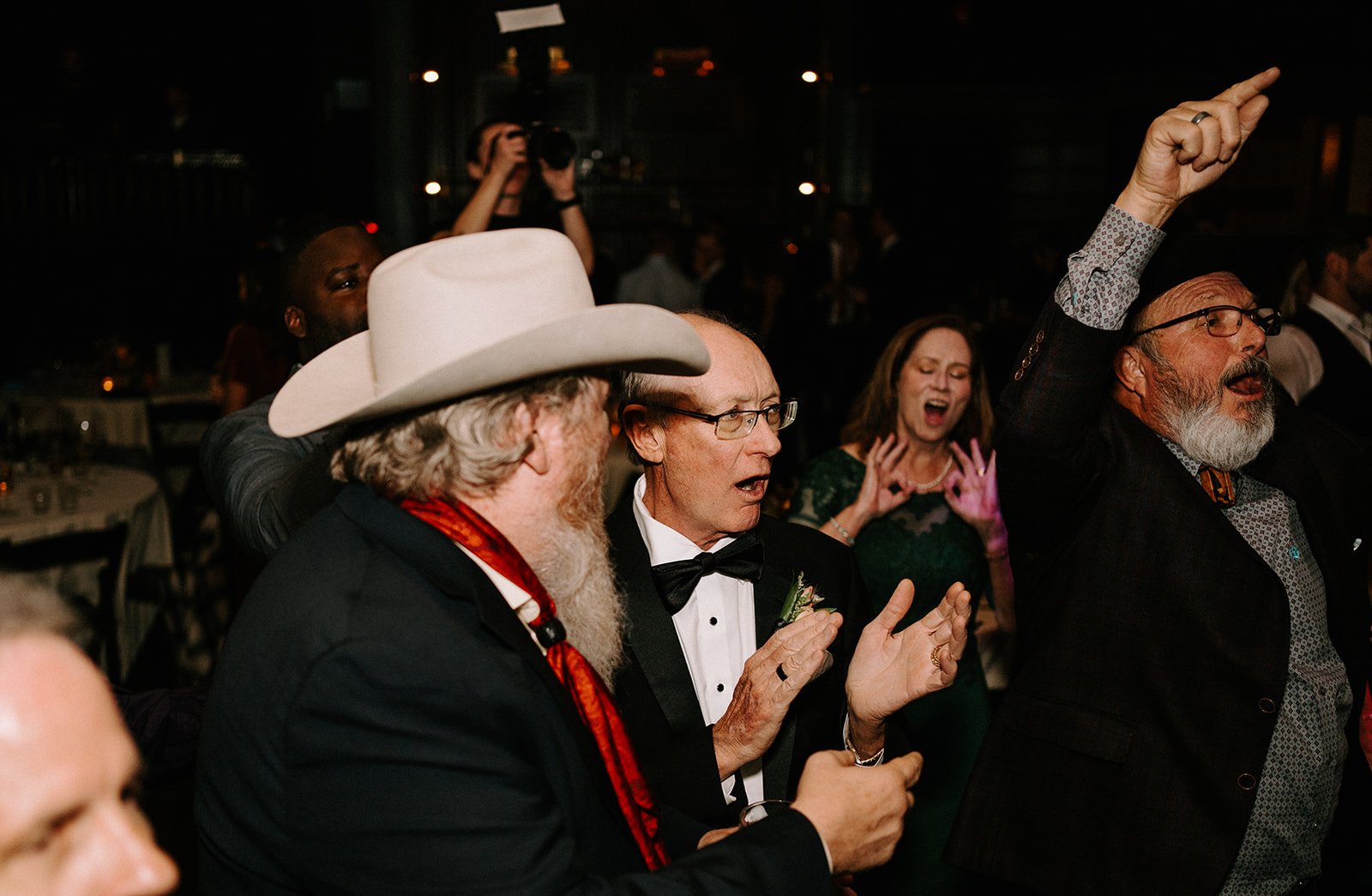 Group of people celebrating at a party, with a man in a cowboy hat, another in a tuxedo clapping, and others cheering enthusiastically.