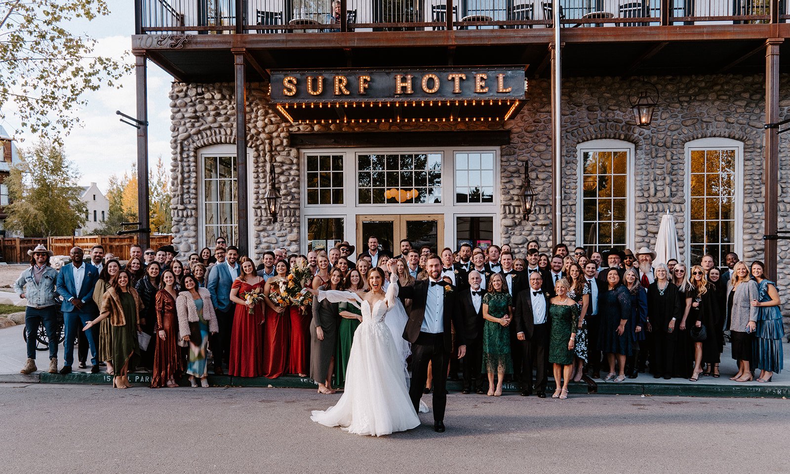 Wedding group photo in front of Surf Hotel, featuring bride in white gown, groom in tuxedo, and guests in formal attire.
