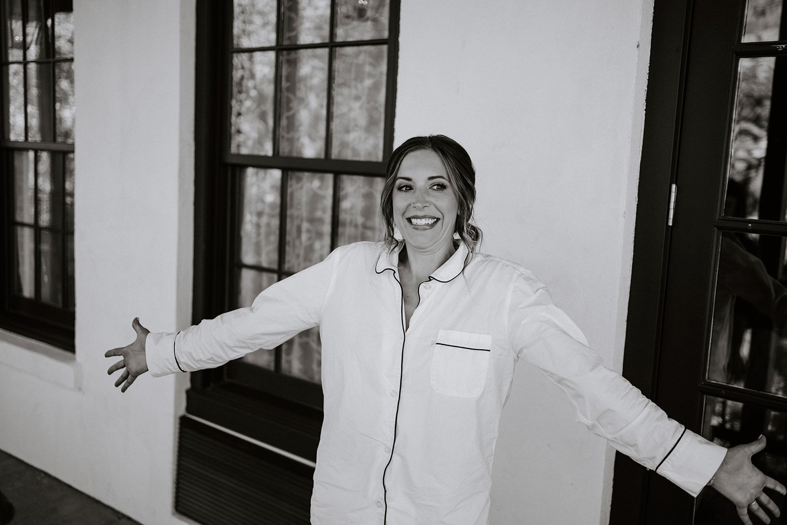 Person smiling, wearing a white shirt with black piping, arms outstretched, standing indoors near a window and door.