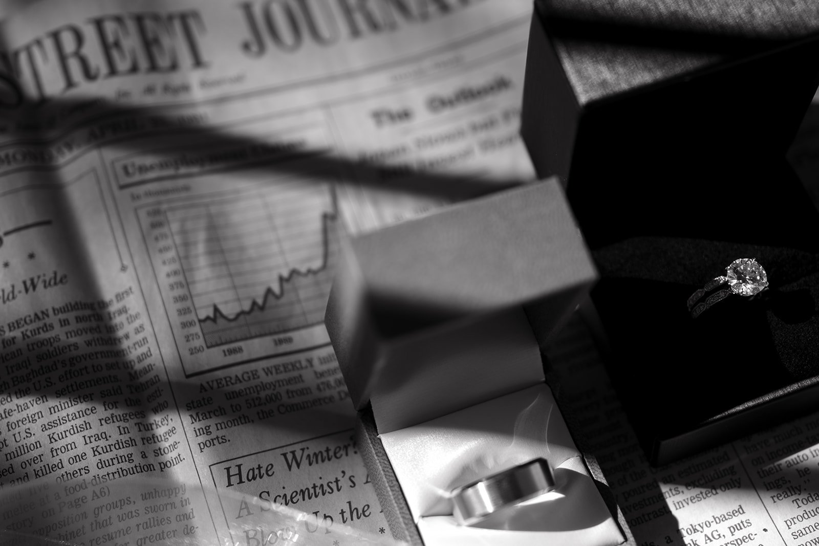 Black and white image showing a newspaper with two open ring boxes, one containing a diamond ring and the other a plain band.