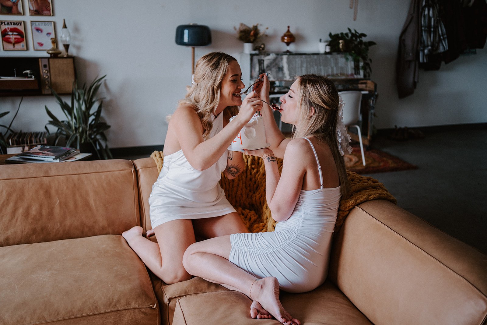 Two women in white dresses sharing a dessert on a couch in a living room.