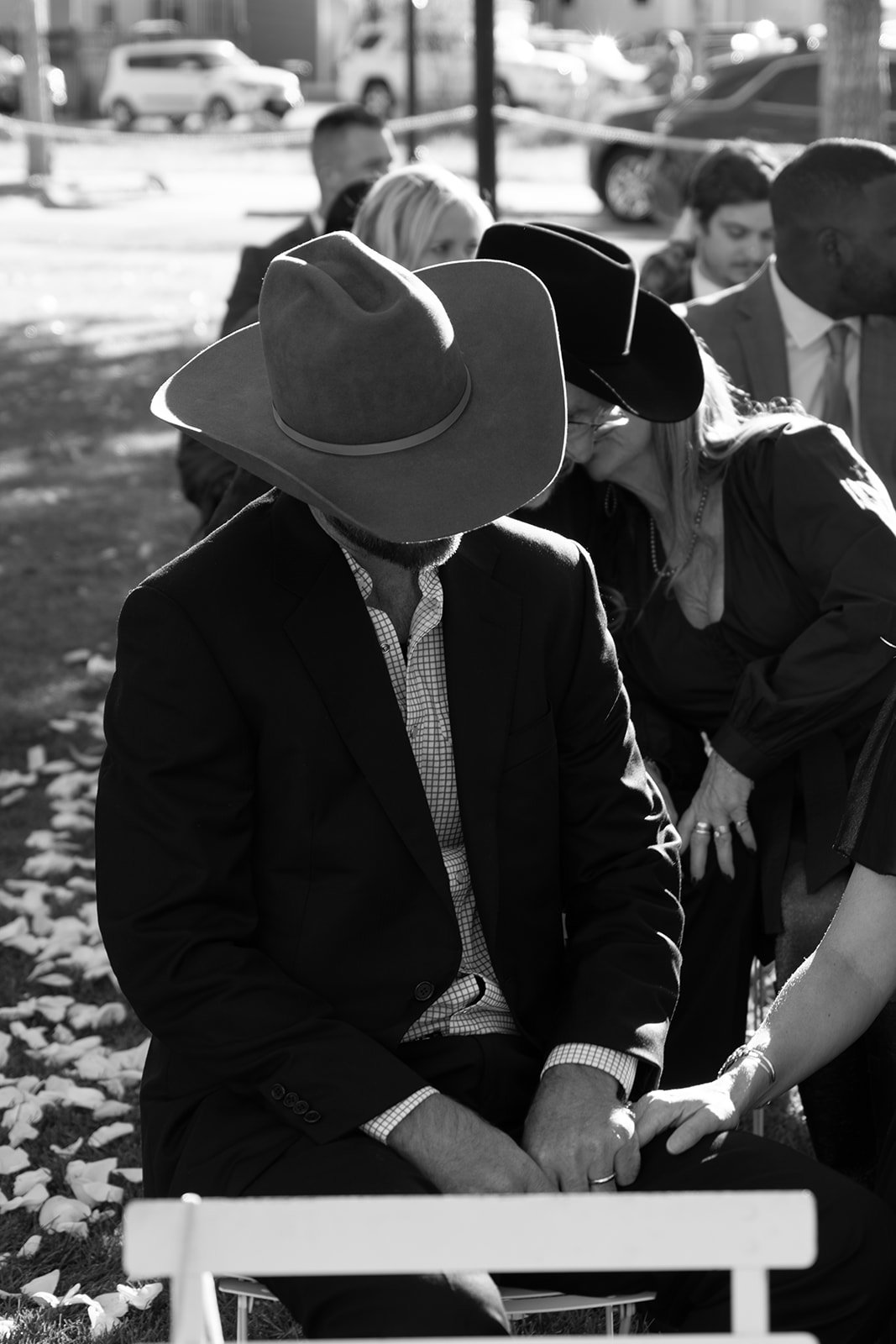 Black and white photo of a man sitting outdoors, wearing a cowboy hat and suit, holding hands with another person. Several people seated nearby, some wearing hats.
