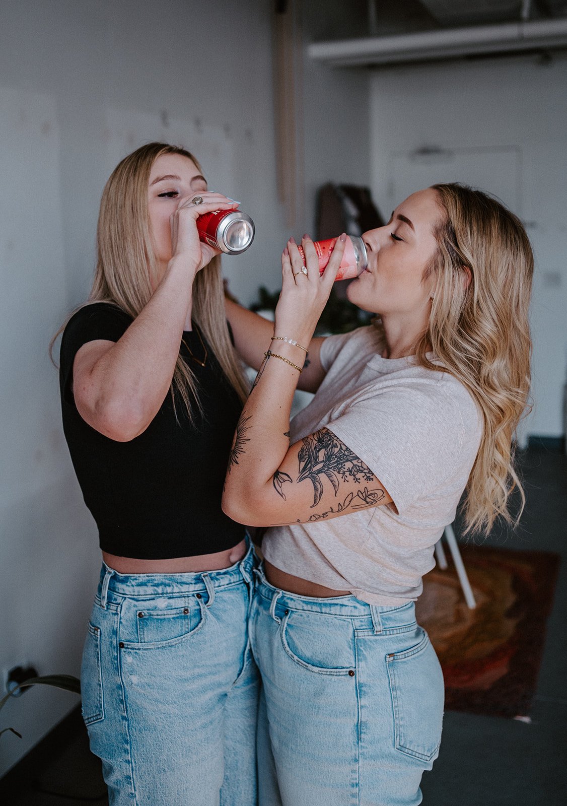 Two women in casual outfits drinking from cans, standing close together with arms around each other, in an indoor setting.