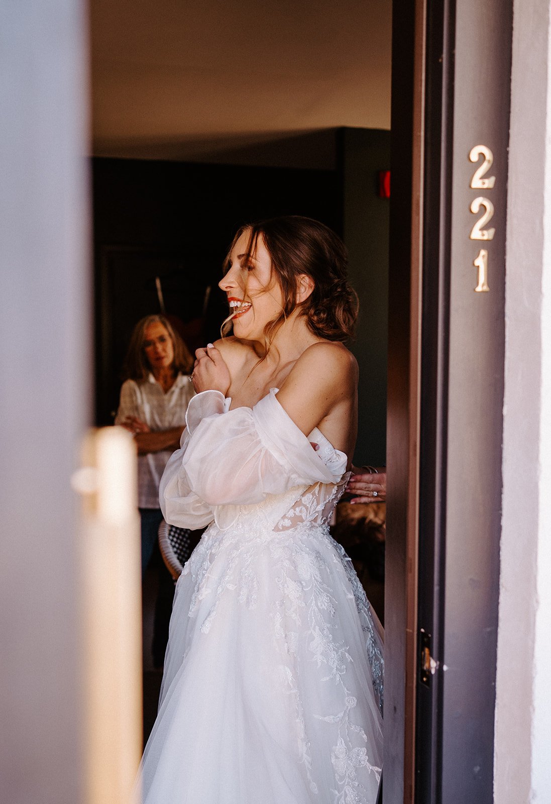 Bride in a white wedding dress, smiling and holding her hands together, with room number 221 on the doorframe.