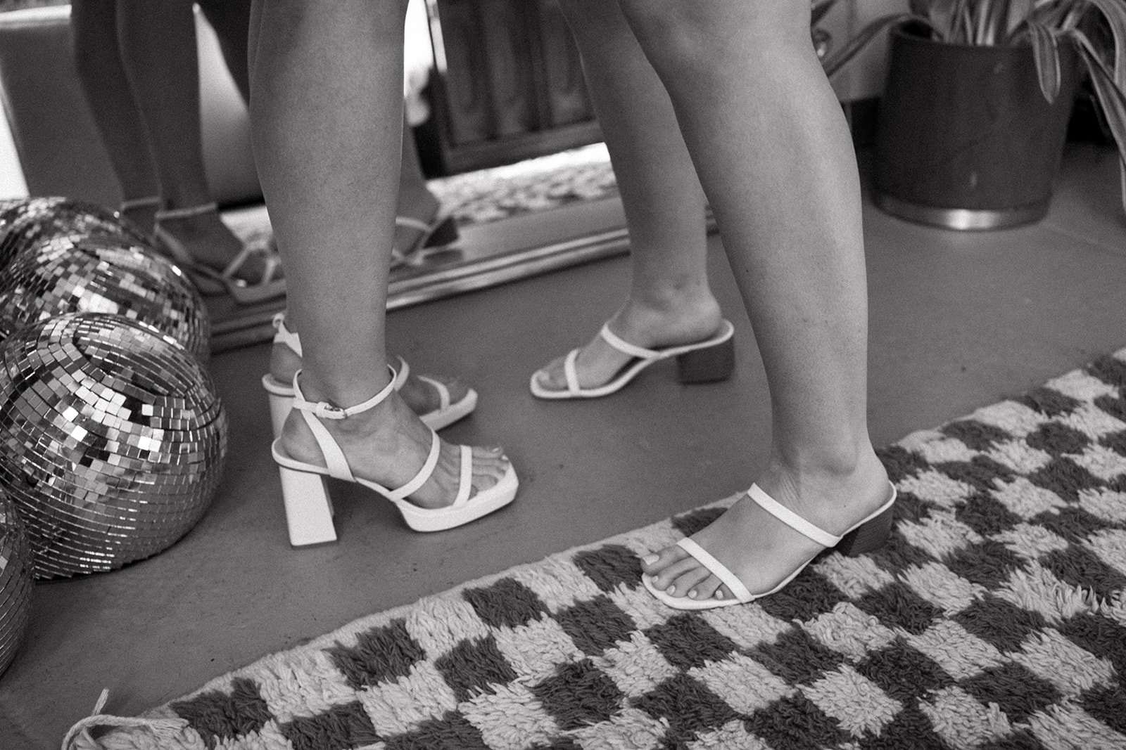 Women wearing strappy heeled sandals standing on a patterned rug with disco balls nearby, in front of a mirror.