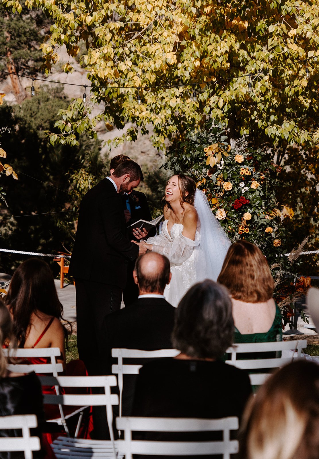 Outdoor wedding ceremony under tree with string lights, bride and groom exchanging vows, guests seated.