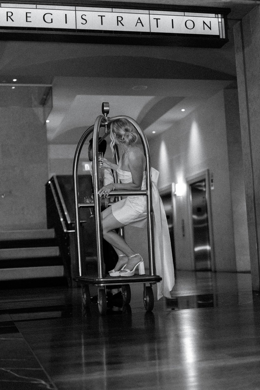 Black and white photo of a woman sitting on a luggage cart under a "Registration" sign, wearing a white dress and holding a drink.
