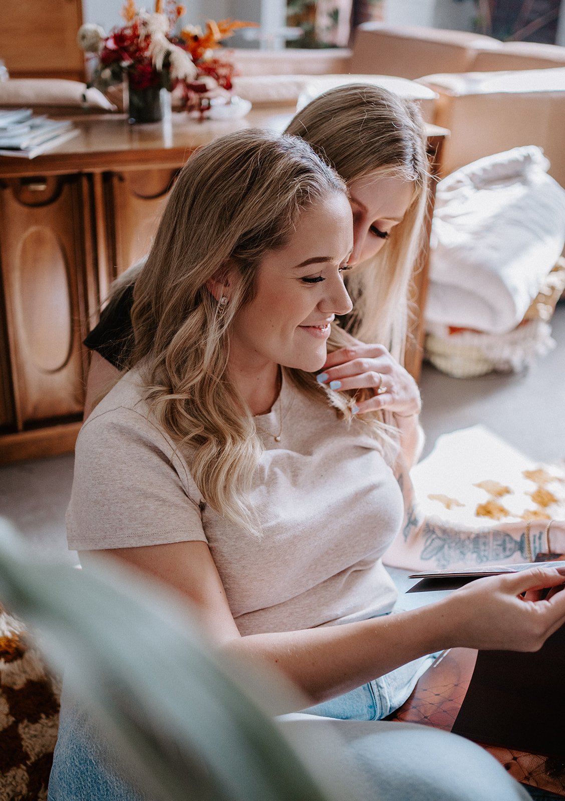 Two women sitting together at home, one embracing the other from behind, smiling, with decorative autumnal flowers on a table in the background.