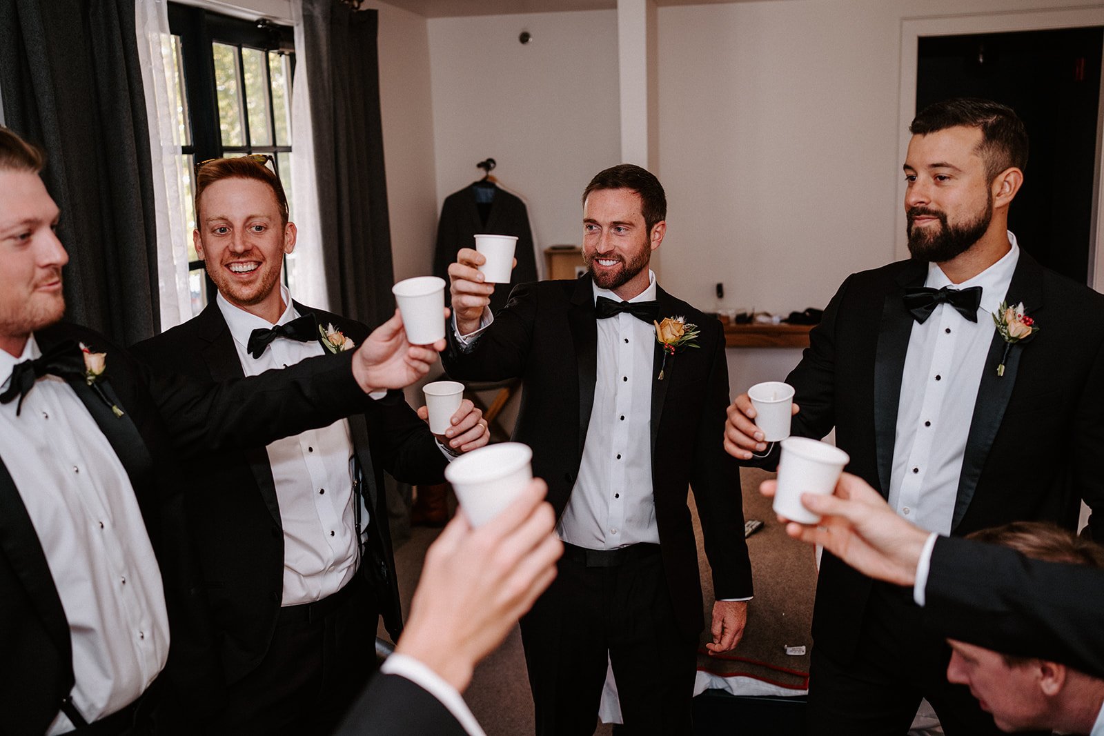 Group of groomsmen in tuxedos toasting with paper cups in a room.