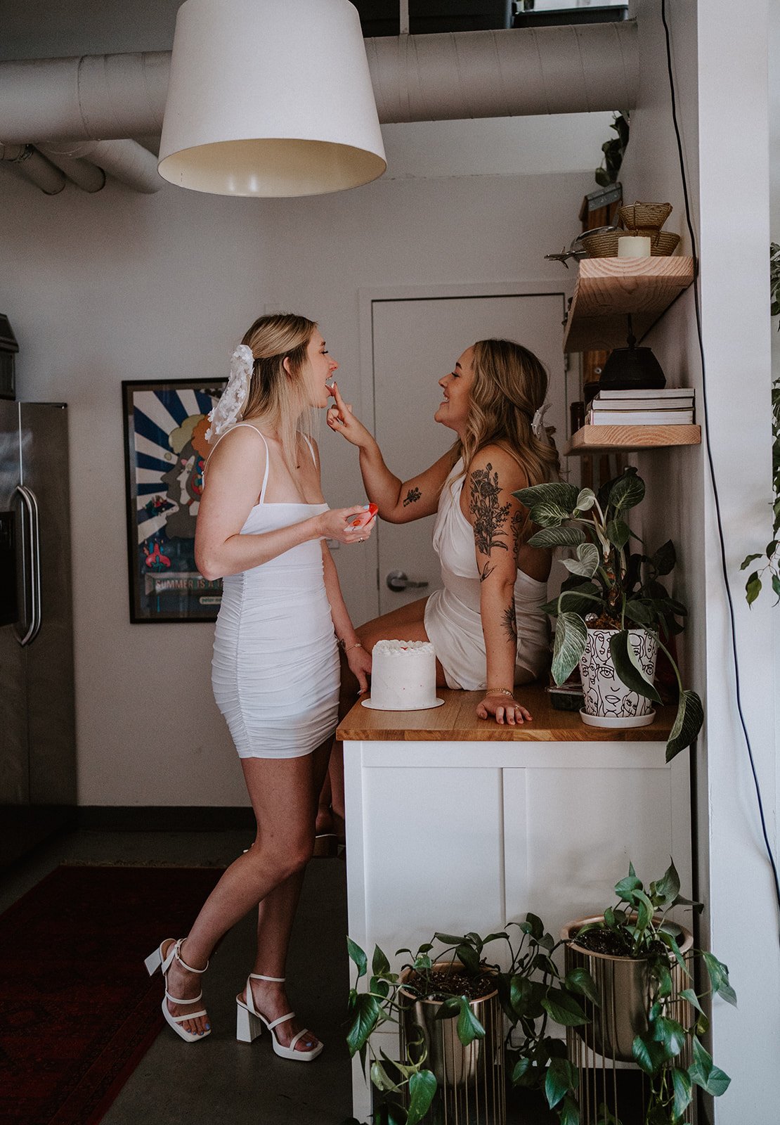 Two women in white dresses enjoying cake in a cozy indoor setting with plants and a modern lamp.