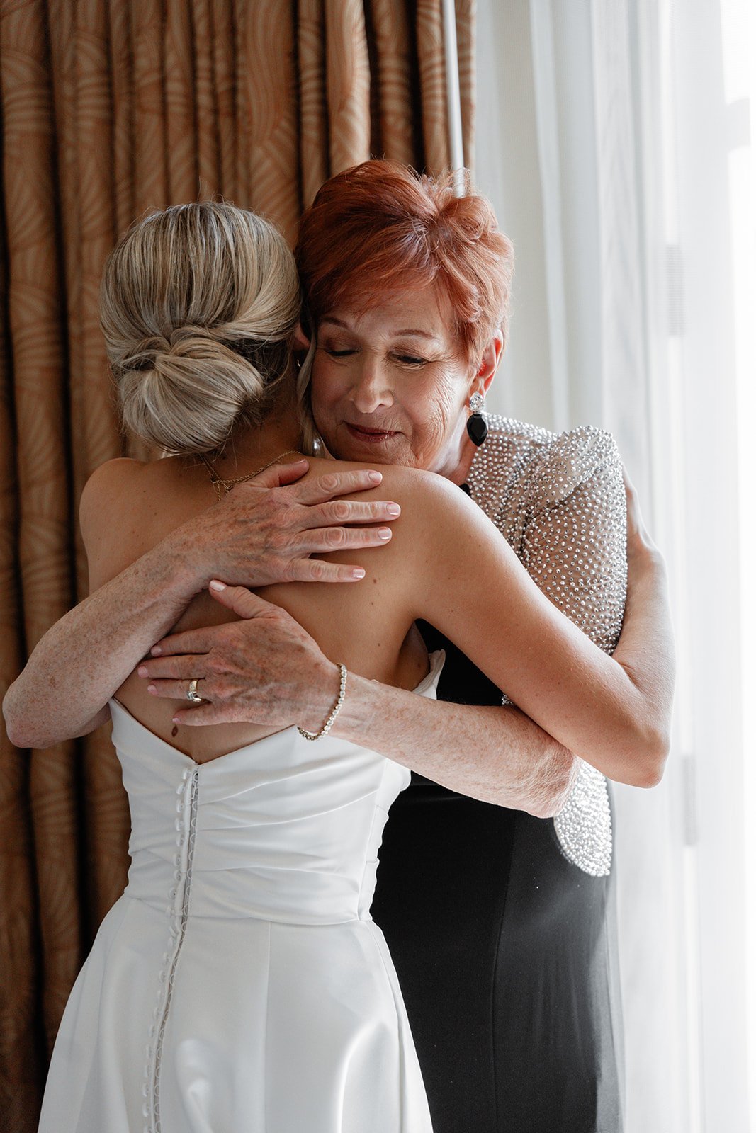 Two women embracing in a warm hug, featuring one in a white dress with a back button detail and the other with short red hair wearing a sparkling gown.