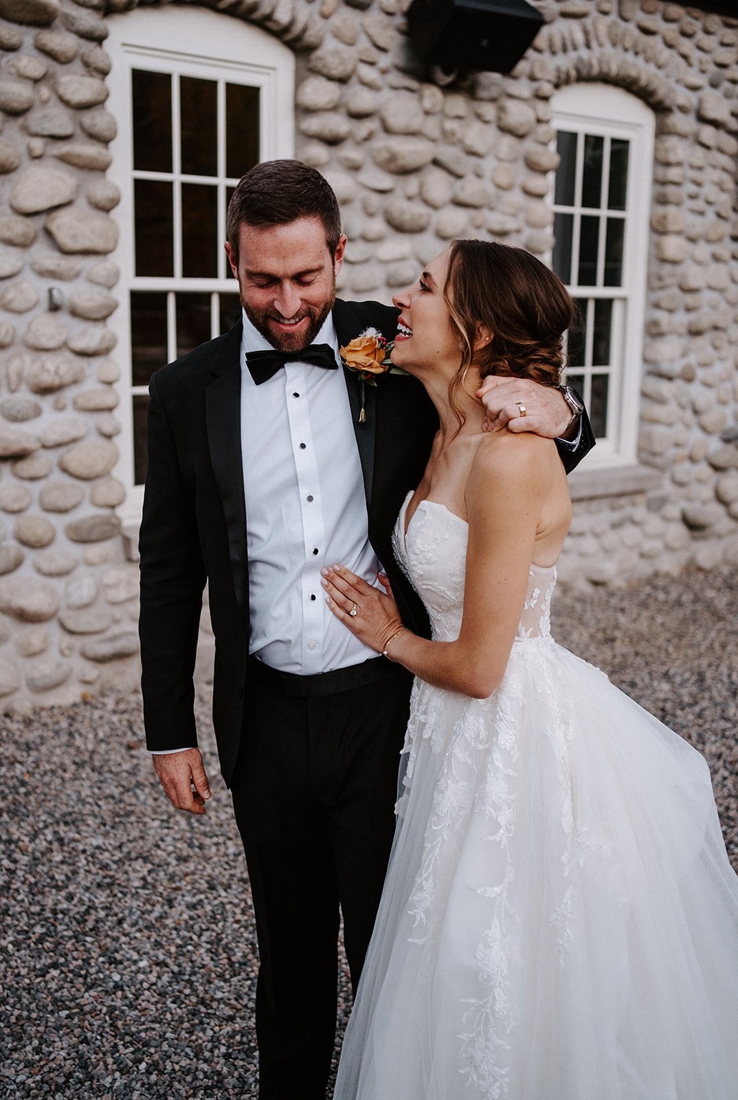 Bride and groom happily embracing outside of a stone building, with the bride wearing a white wedding dress and the groom in a black suit with a bow tie.