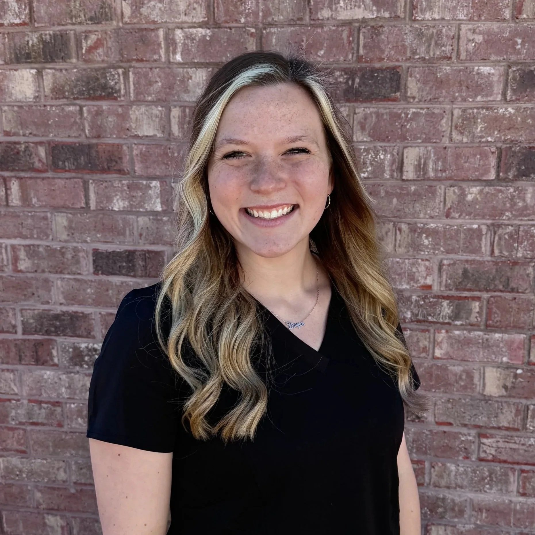 A woman with long wavy blonde hair, wearing a black top, smiling and standing in front of a red brick wall.