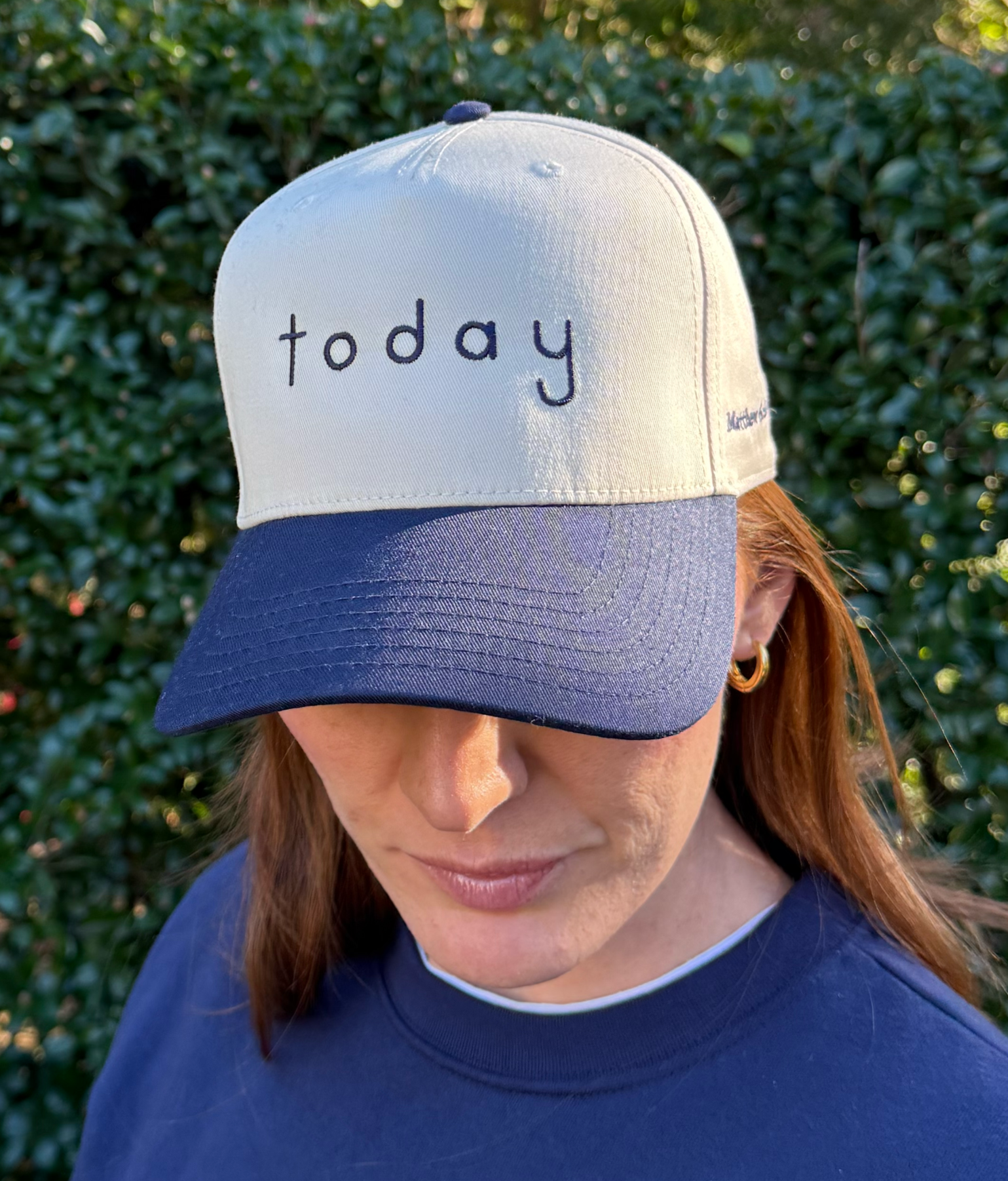 A woman with reddish hair wearing a navy and white cap with the word 'today' embroidered on it, standing outdoors with green foliage in the background.
