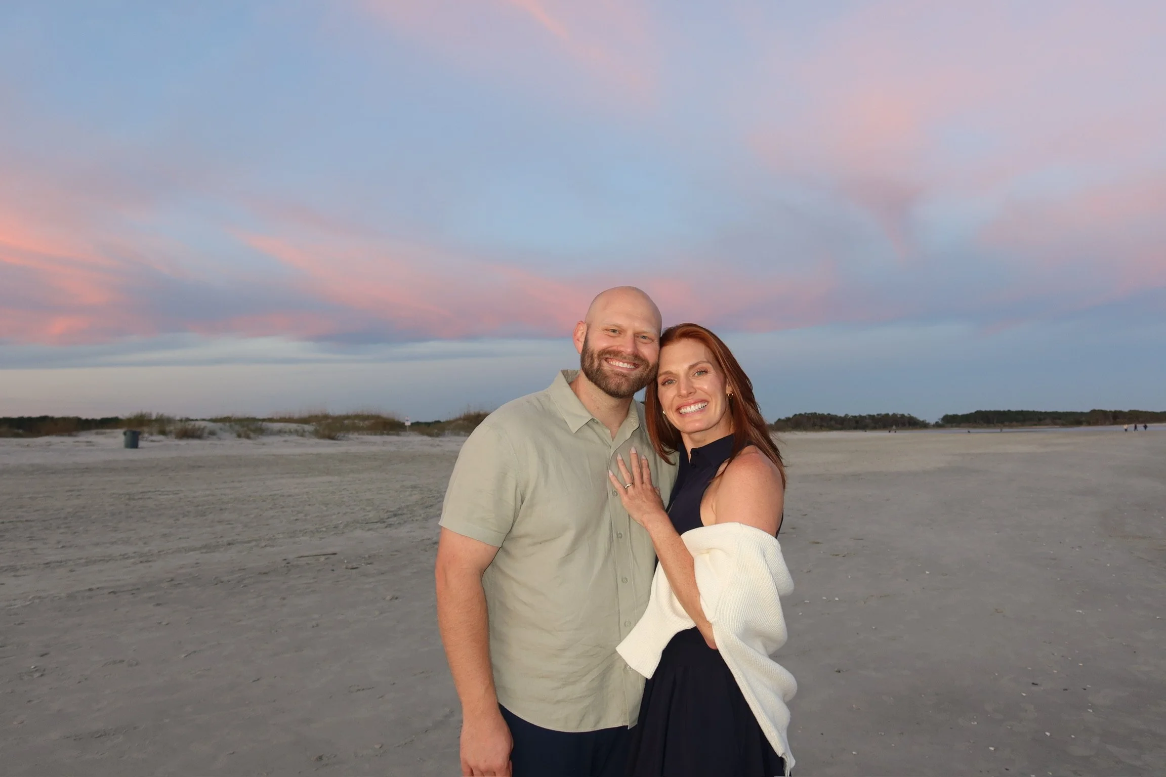 A smiling couple stands together on a beach at sunset, with pink and blue sky in the background.