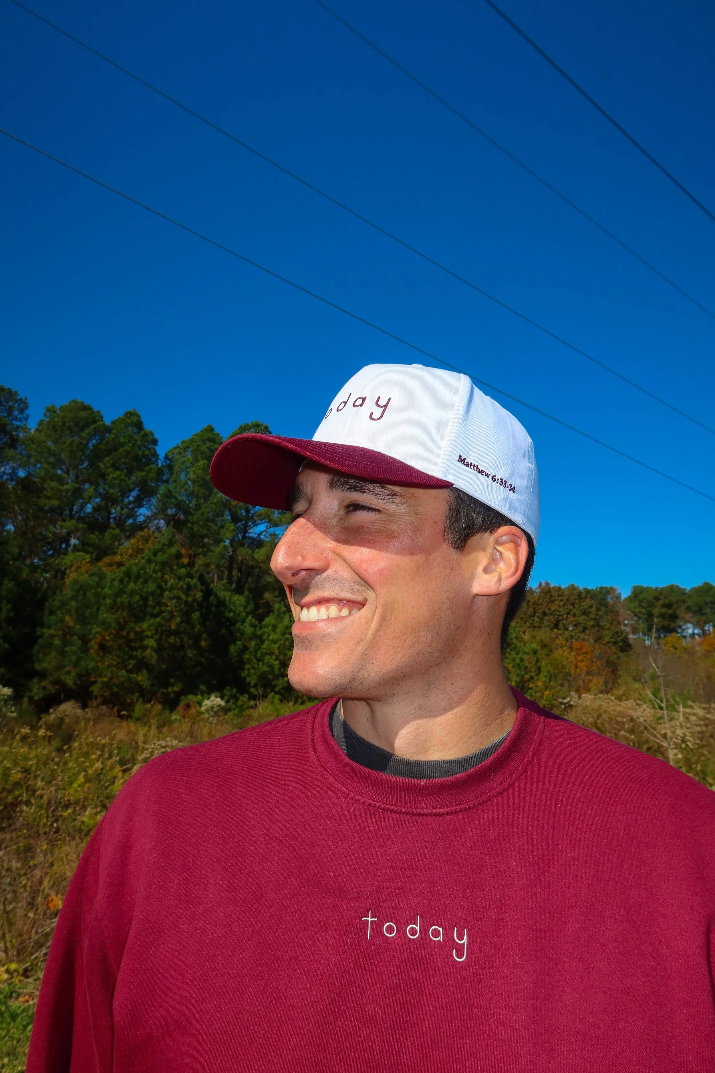 A smiling man outdoors wearing a maroon sweatshirt and a white baseball cap that says 'today' in red. The sky is blue with power lines overhead and trees in the background.