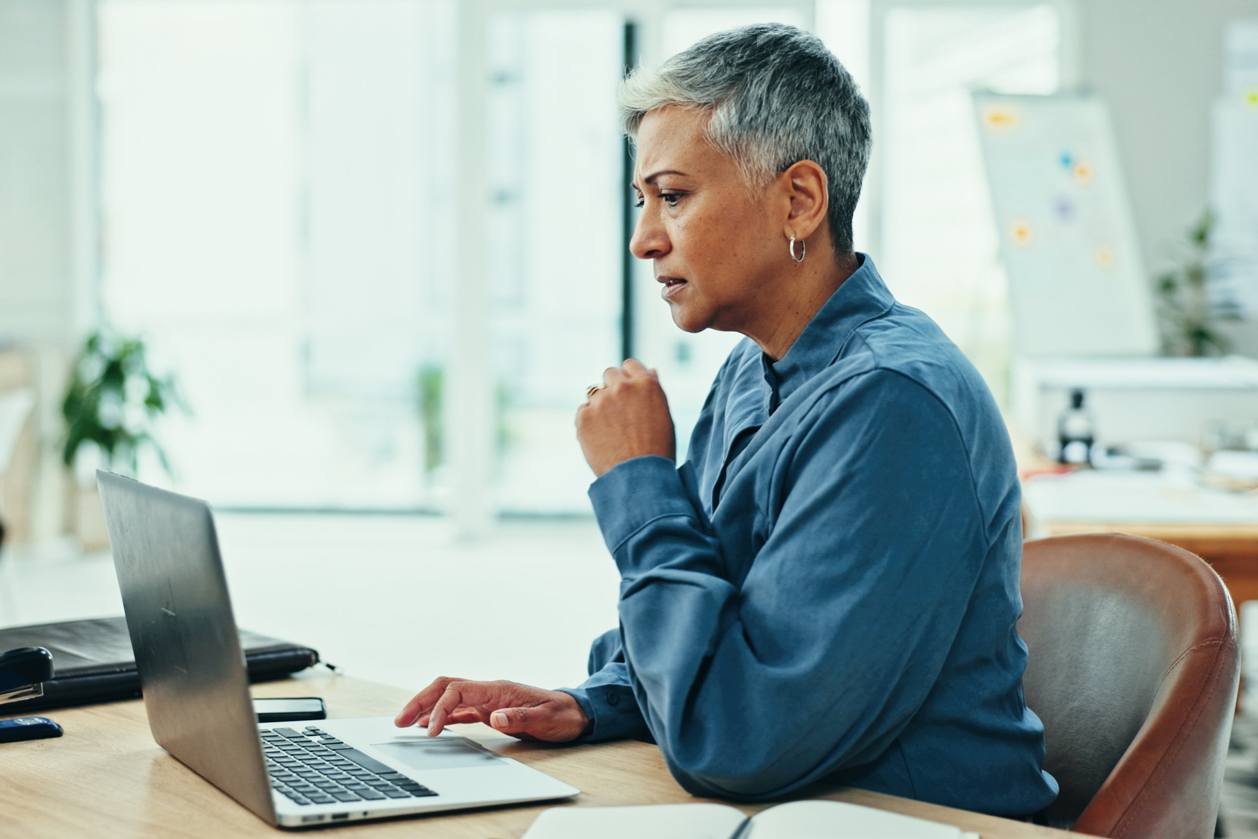 Older woman with short gray hair and hoop earrings working on a laptop at a wooden table in a bright, modern office. She appears focused and is wearing a blue shirt.