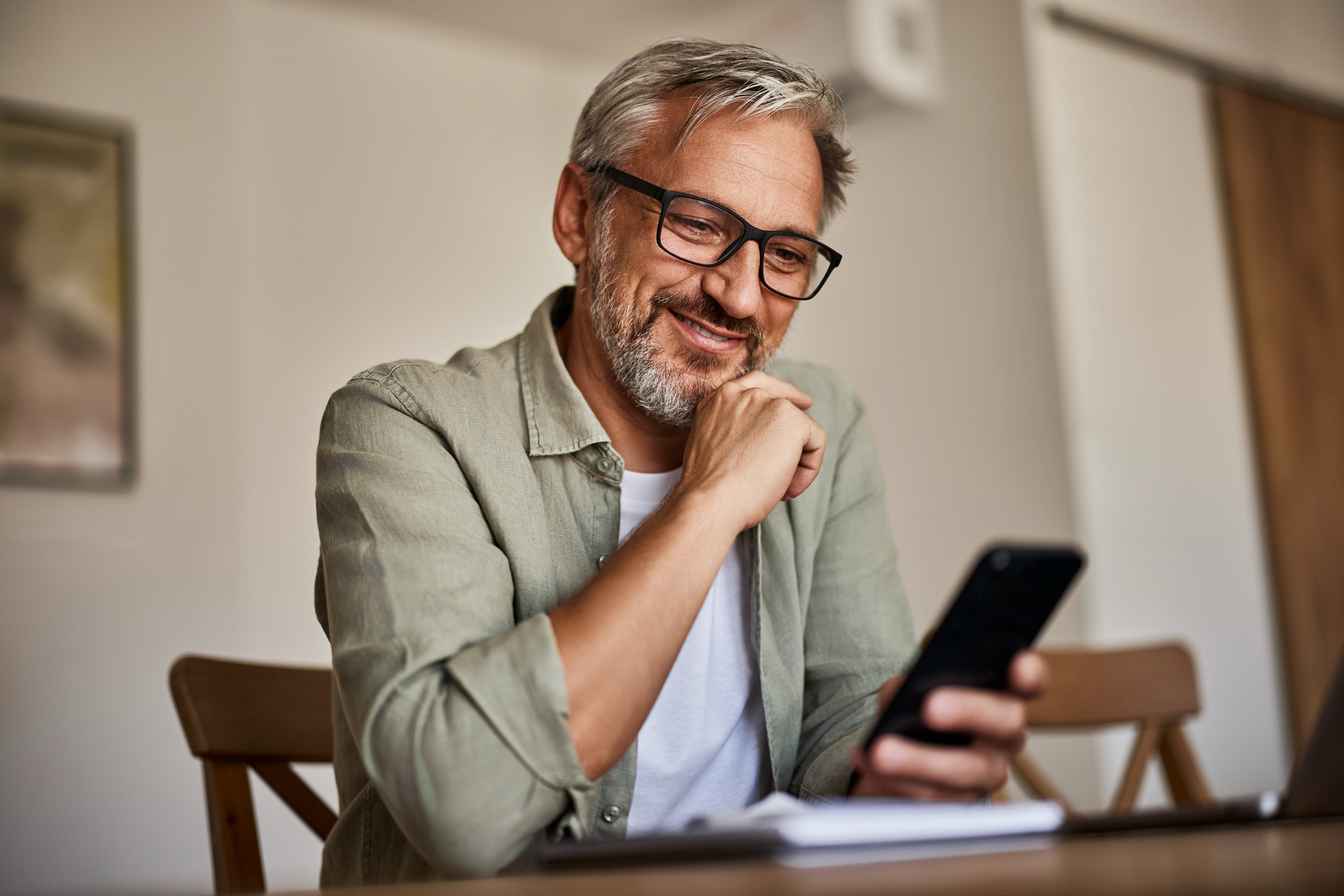 An older man with gray hair, beard, and glasses sitting at a wooden table, smiling as he looks at his phone in a room with neutral-colored walls and artwork.