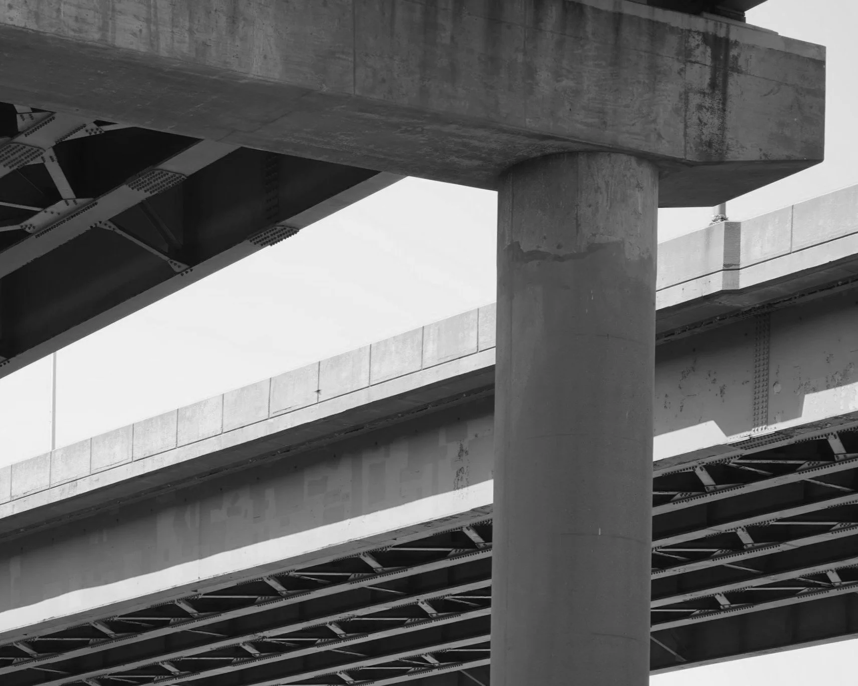 Black and white photo of a concrete bridge structure showing a large cylindrical support pillar and the underside of the bridge with its metal framework.