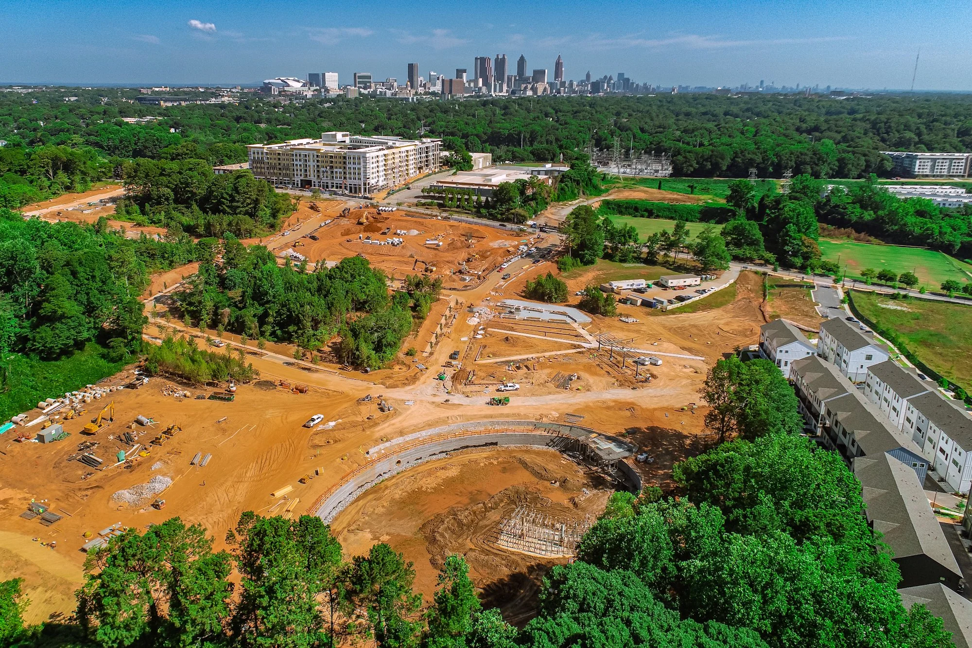 Construction site with cleared and excavated land, dirt roads, and construction equipment, adjacent to a paved road and surrounded by trees.