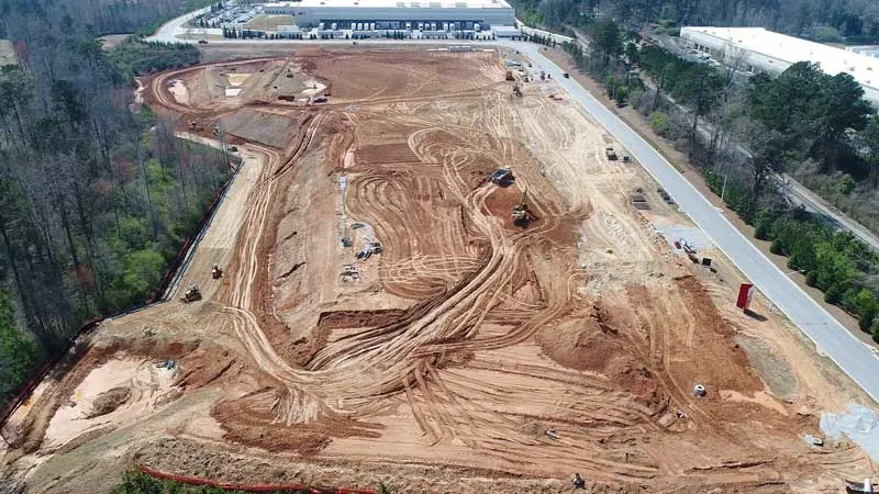 Construction site with cleared and excavated land, dirt roads, and construction equipment, adjacent to a paved road and surrounded by trees.