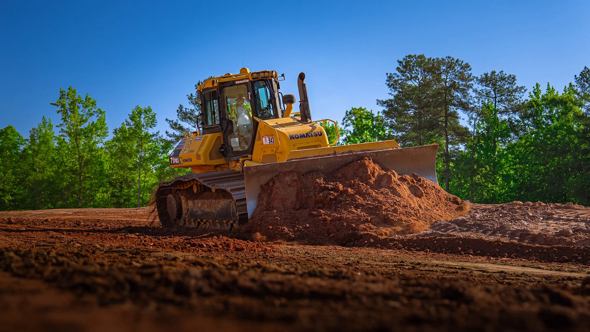 Aerial view of a large construction site with dirt tracks, construction vehicles, and partially built structures, adjacent to a paved road and trees.