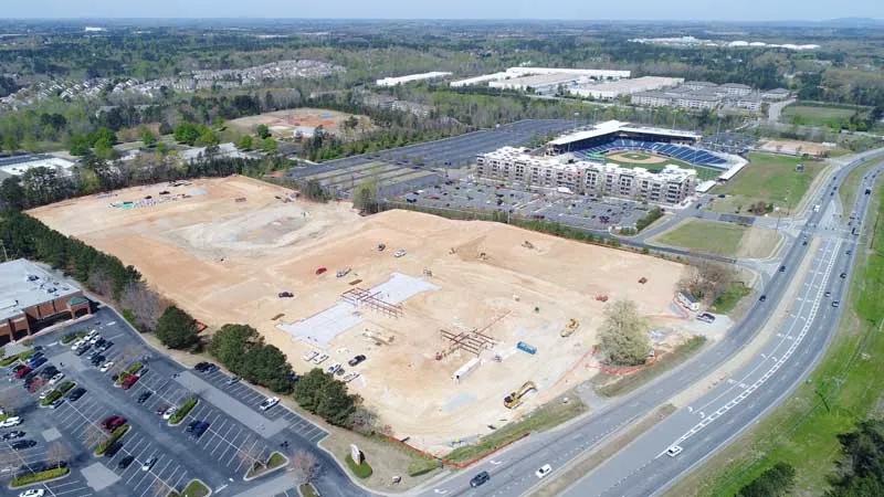 Construction site with cleared land, parking lots, and a stadium in the background.