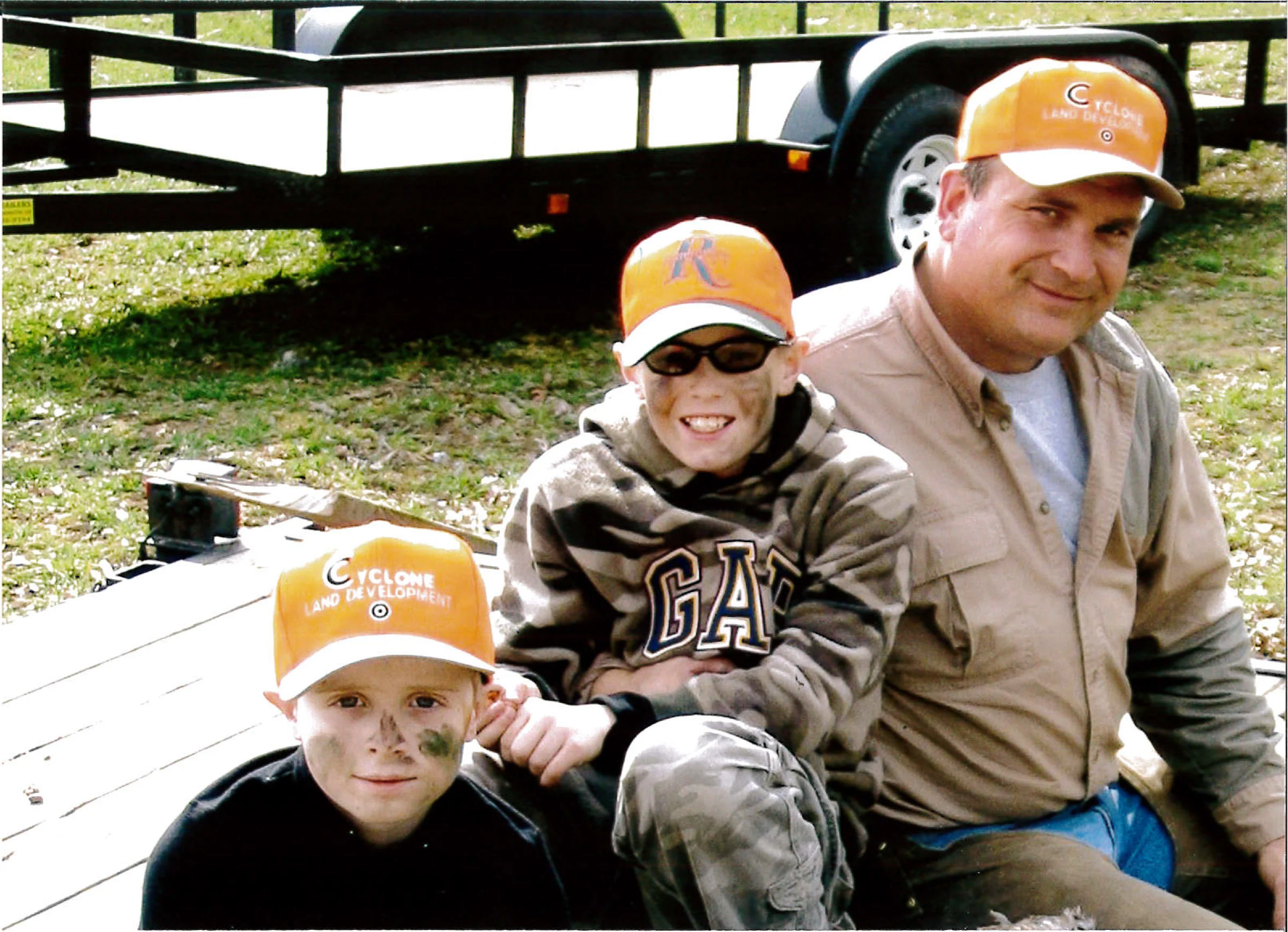 Three children and an adult sitting on a wooden surface with a trailer behind them outdoors. All are wearing orange hats, with two of the children having face paint and camouflage clothing.