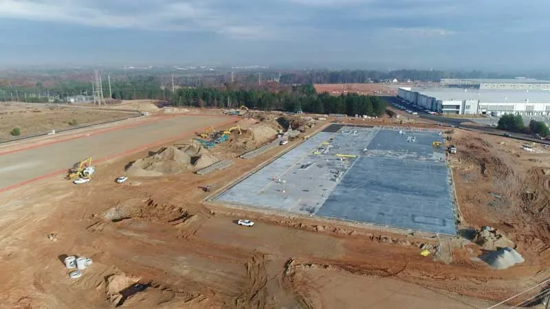Construction site with a large concrete foundation, construction vehicles, and dirt piles, with industrial buildings in the background.