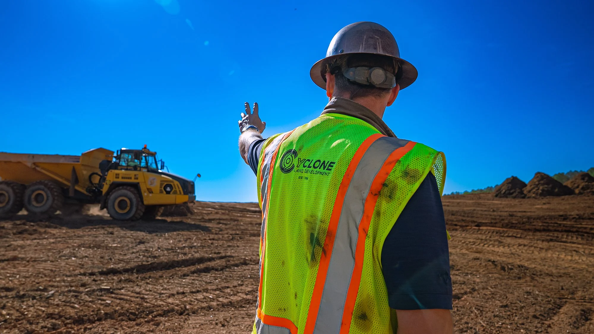 Aerial view of a construction site with excavators working on reddish soil, surrounded by a forest in the background.