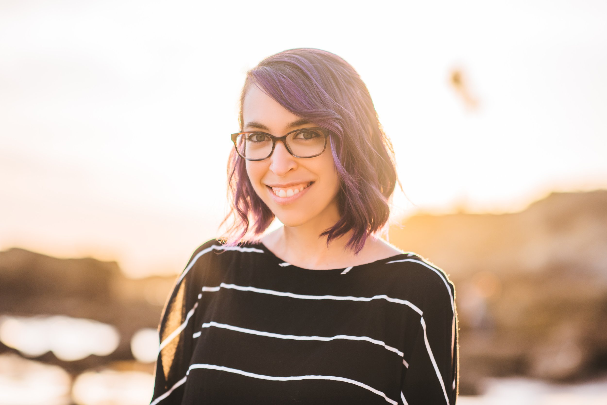 Young woman with purple hair and glasses smiling outdoors during sunset.